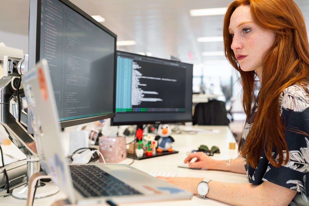 service-05 A focused female software engineer coding on dual monitors in a modern office.