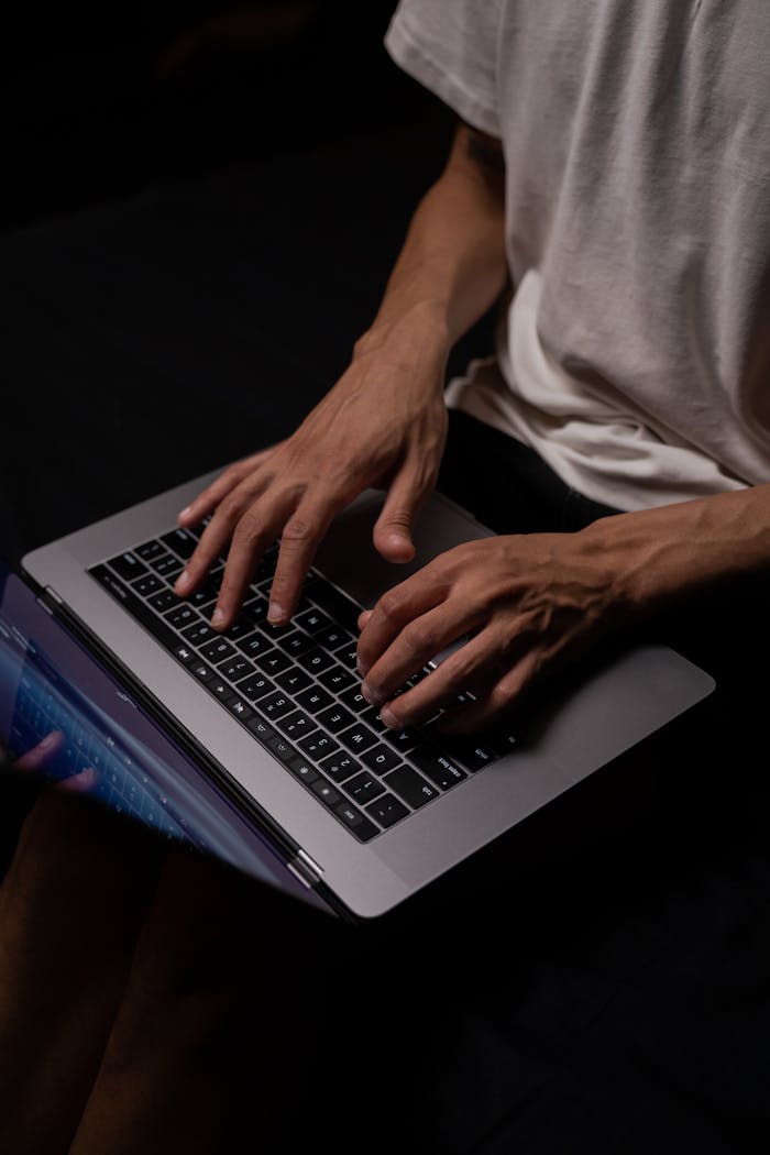 An adult male programmer typing on a laptop in a dimly lit room, concentrating intently.